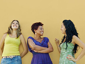 Drei Frauen stehen vor einer gelben Wand, zwei Frauen unterhalten sich, eine Frau schaut nach oben Richtung Himmel 