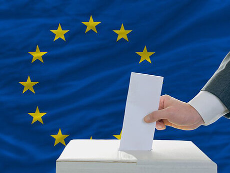 Person wirft vor der EU-Flagge einen Wahlzettel in eine Wahlurne
