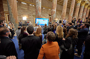 Gruppe von Menschen schaut in Säulenhalle des Parlaments auf Bildschirm mit vorläufigem Wahlergebnis der NR-Wahl 2024