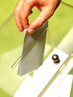 Eine Hand steckt einen zusammengefalteten Stimmzettel in eine durchsichtige Wahlurne.