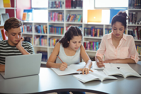 Jugendliche lernen in einer Bibliothek, vor ihnen Bücher und ein Laptop